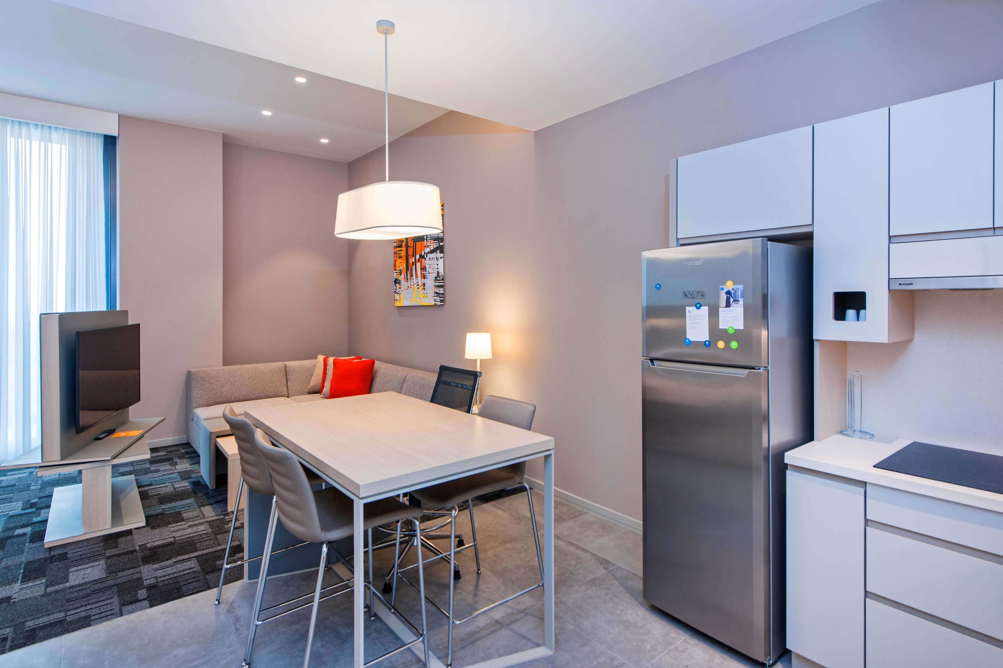 Una habitación de hotel moderna con comedor, sofá con cojines rojos, refrigerador grande y televisión.