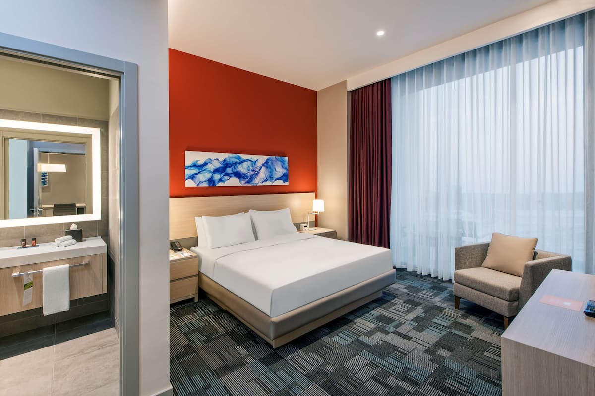 A hotel room with a bed, a desk, a chair, a bathroom with a mirror, and a large window with curtains.