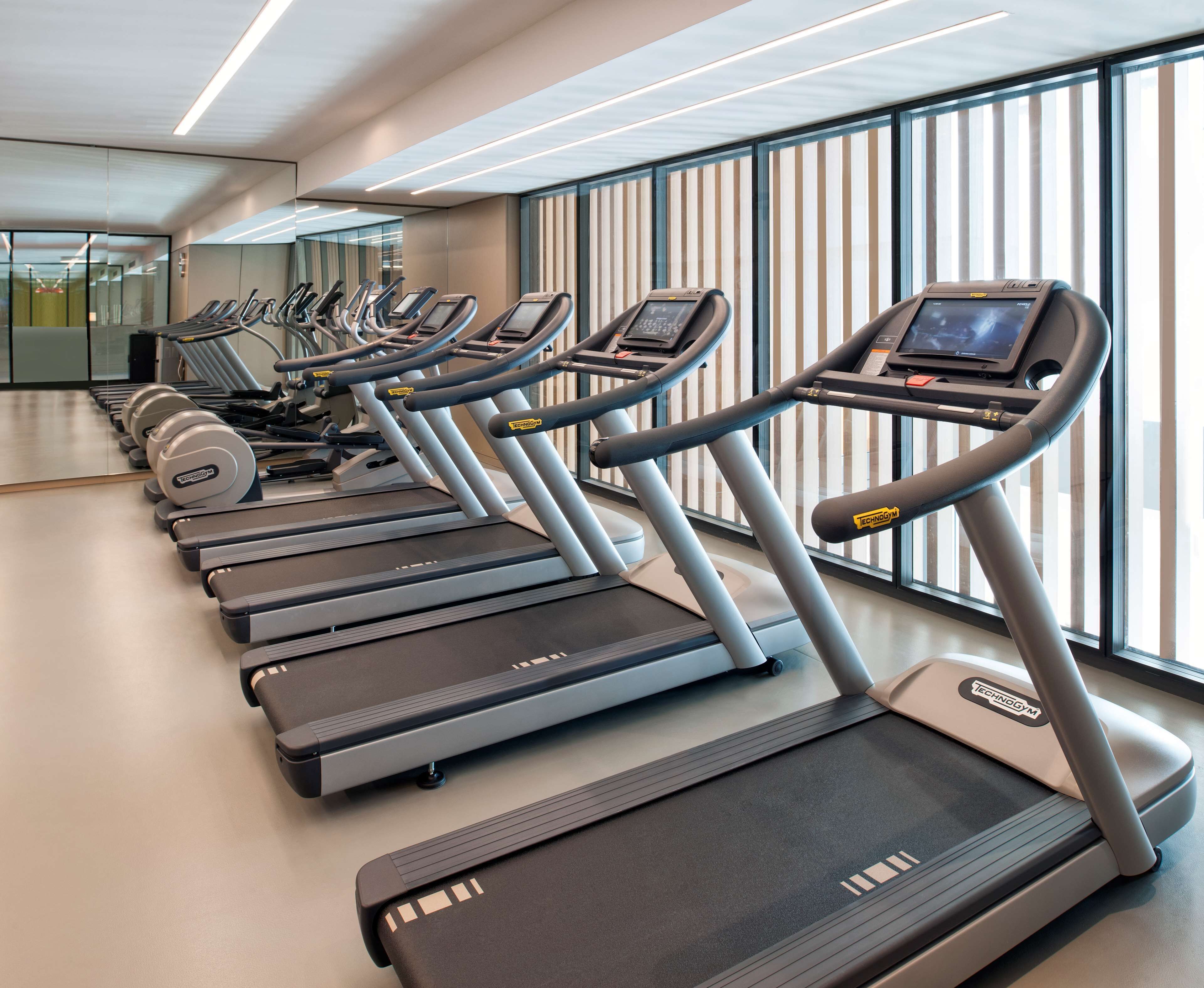 Eine Reihe von Laufbändern in einem Fitnessstudio mit großen Fenstern und modernem Design.