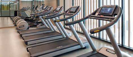 Eine Reihe von Laufbändern in einem Fitnessstudio mit großen Fenstern und modernem Design.
