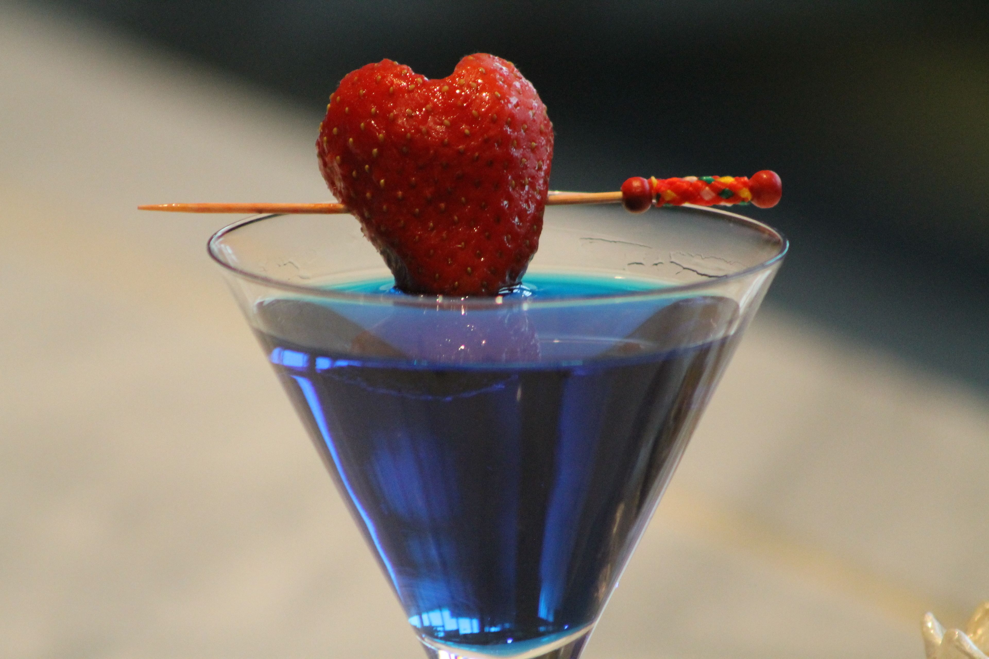 A cocktail with a heart-shaped strawberry on a skewer in a martini glass filled with blue liquid.