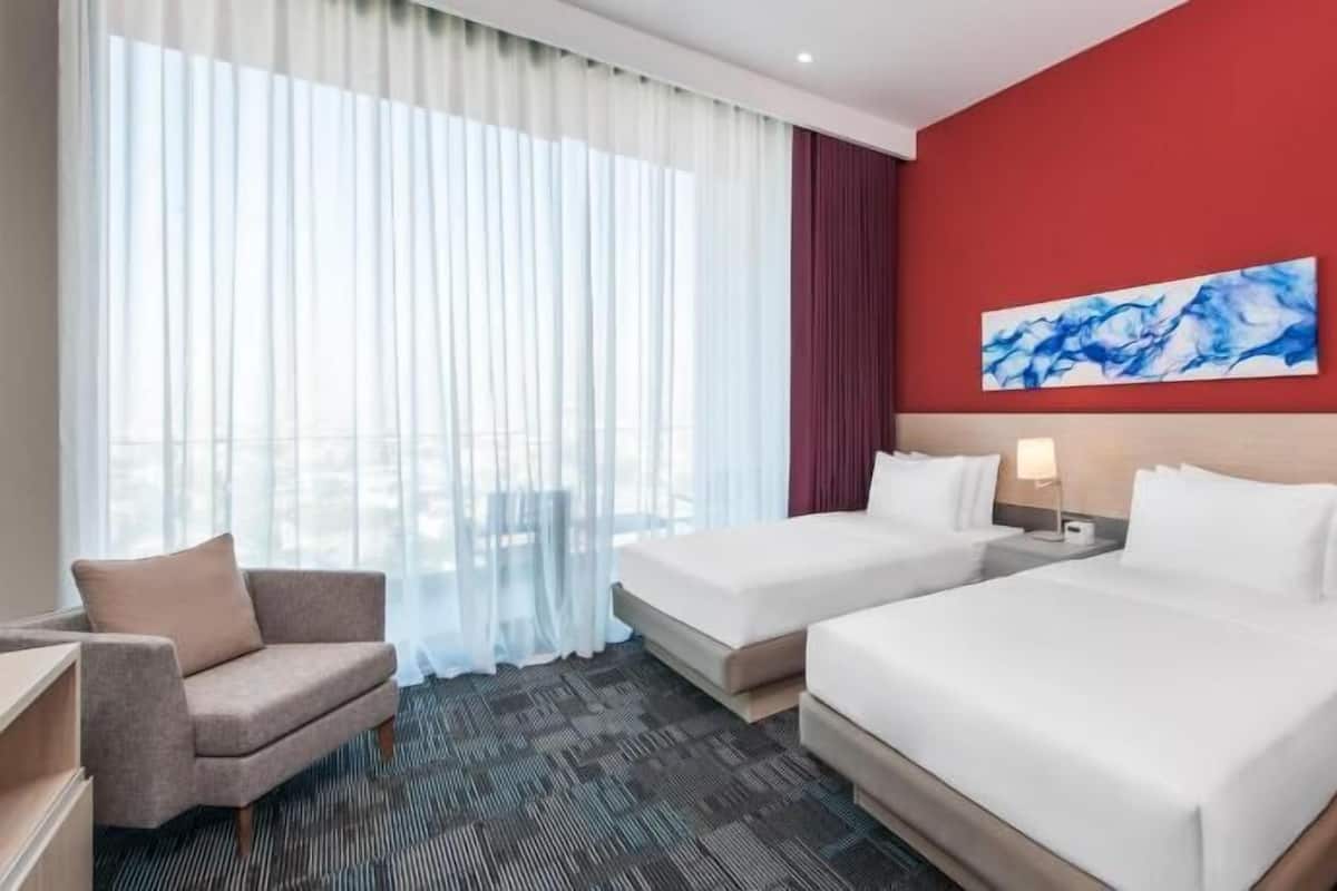 A hotel room with two beds, a sofa, a desk, and a large window with sheer curtains.