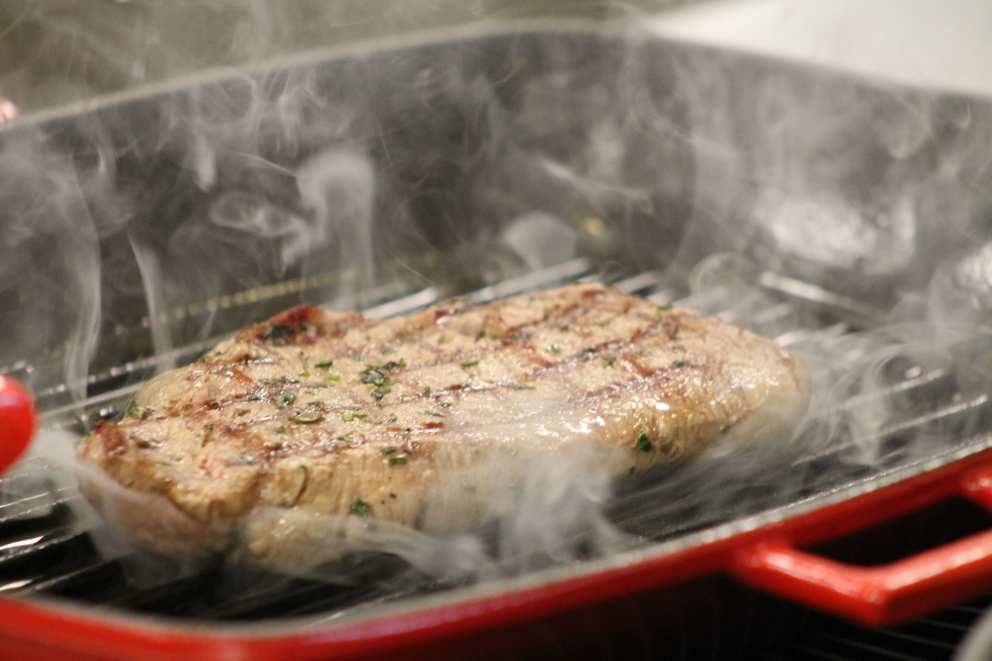 A steak being grilled on a red grill pan with visible smoke.