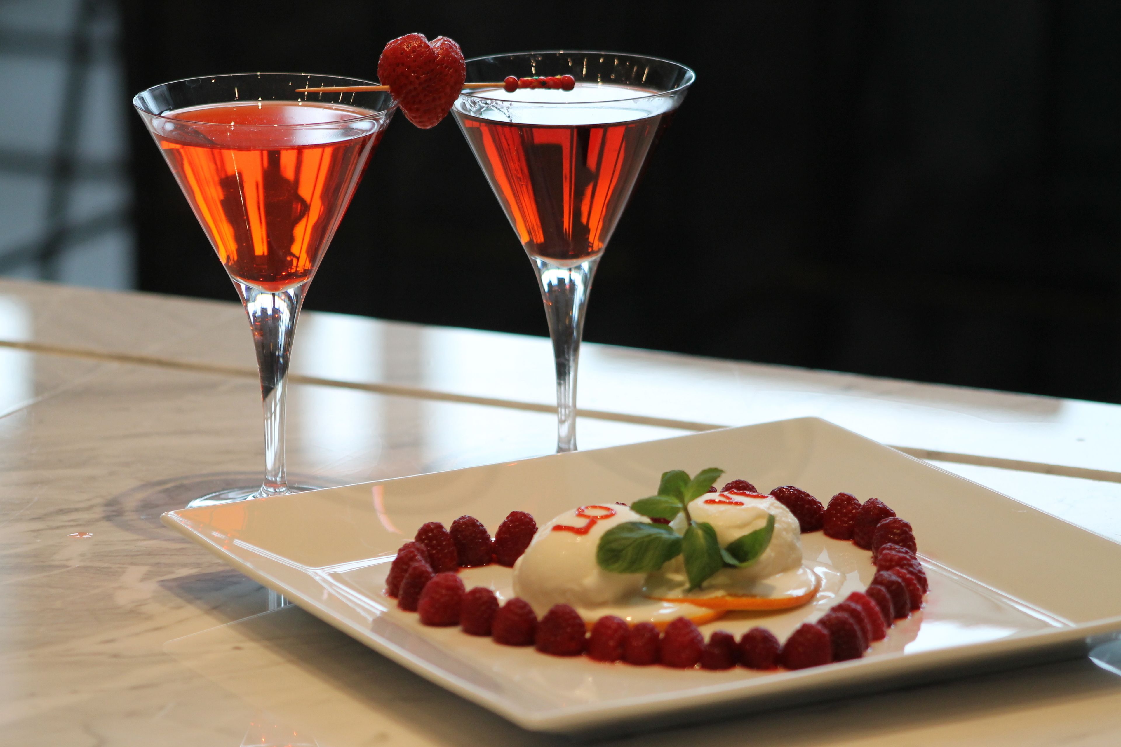Dos copas de martini con una bebida roja y un plato de postre con frambuesas y una bola de helado.