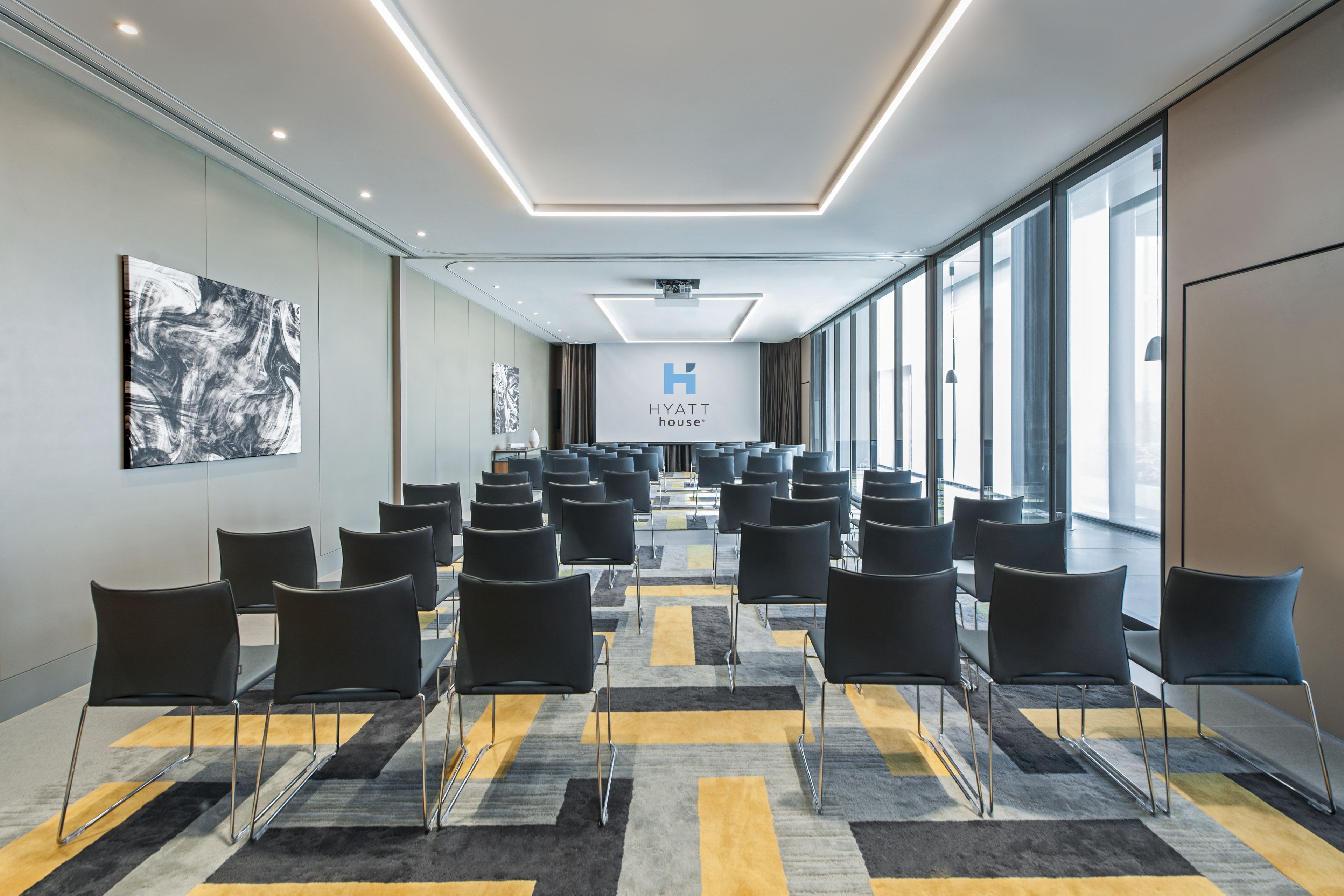 A modern conference room with rows of black chairs, a large screen displaying the Hyatt House logo, and large windows providing natural light.