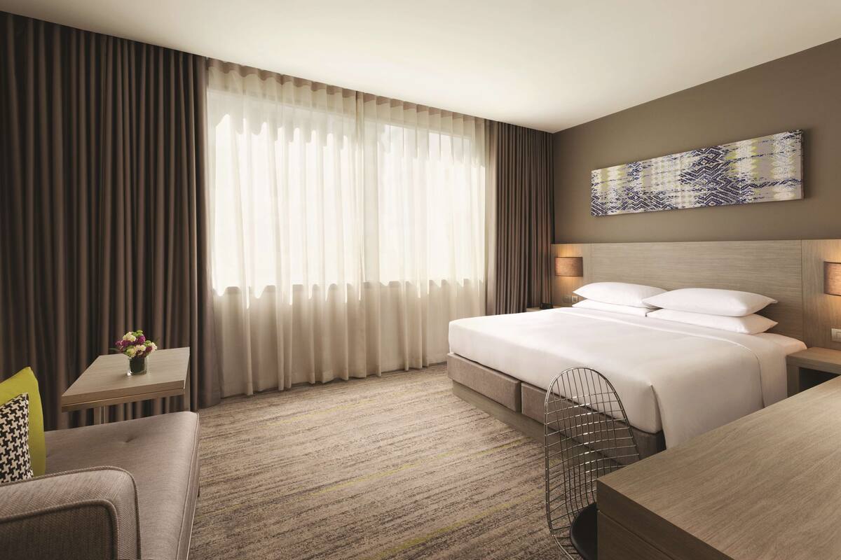 Une chambre d’hôtel avec un grand lit, un bureau, une chaise, une petite table ornée d’un vase de fleurs et une grande fenêtre avec des rideaux.