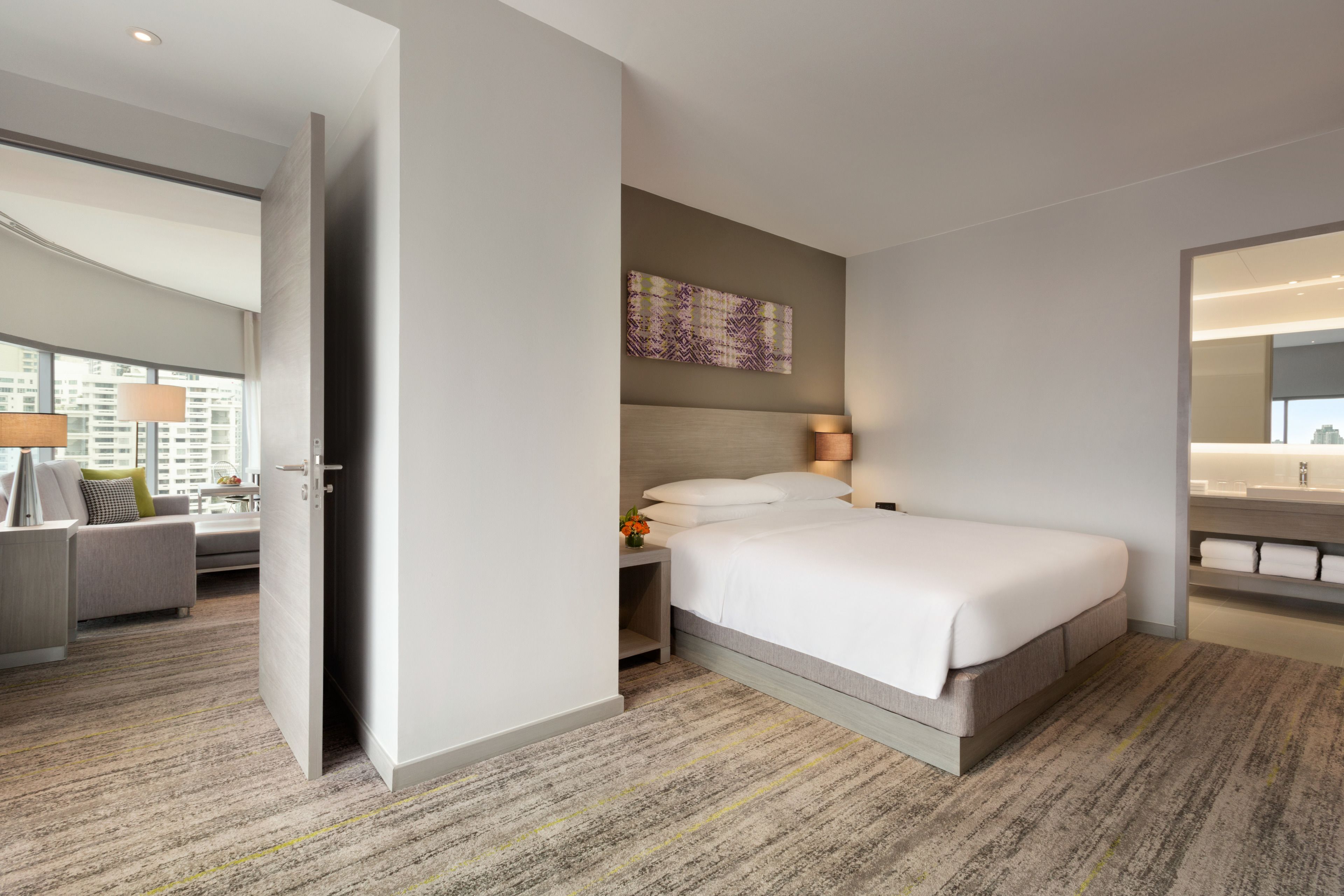Een moderne hotelkamer met een groot bed, een nachtkastje met een lamp, een klein bijzettafeltje met een vaas en uitzicht op de stad door grote ramen.