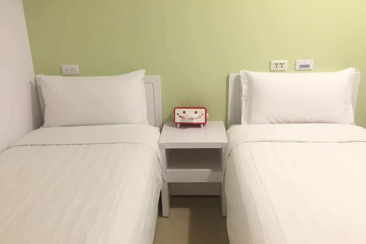 A room with two single beds, a nightstand between them, and a small red alarm clock on the nightstand.