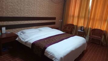Habitación de hotel con una cama grande, cabecera de madera, una mesita de noche con lámpara, una silla y una ventana con cortinas.
