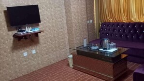 Una habitación con una televisión de pantalla plana montada en la pared, un pequeño estante de madera debajo, una mesa de centro de vidrio con una tetera y tazas, un sofá morado y un piso con diseño de ladrillo.
