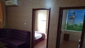 Una habitación con un sofá púrpura, una cama con colcha blanca y morada, un pequeño armario de madera y una puerta que da a otra habitación.