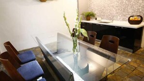 Uma área de jantar moderna com mesa de vidro, cadeiras azuis acolchoadas, um vaso com flores, um painel decorativo de madeira e uma bancada de cozinha com uma fruteira.