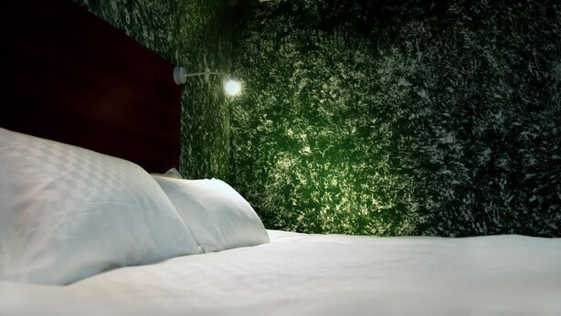 Cama com roupa de cama branca num quarto com parede verde texturizada e cabeceira de madeira escura.