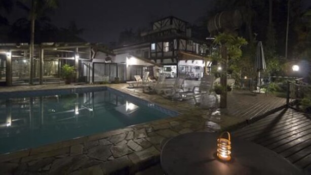 A nighttime view of a pool area with a wooden deck, lounge chairs, and a table with a lantern. - Hotel Britannia (Itatiaia)