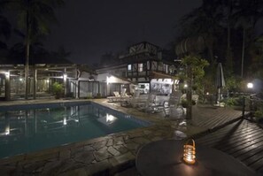 Vista nocturna de un área de piscina con terraza de madera, sillas de descanso y una mesa con farol.
