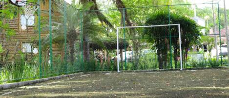 A fenced outdoor basketball court with a goal post, surrounded by trees and a brick building.