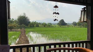 Vista desde una terraza de madera con un campo de arroz, un sendero y árboles, una decoración colgante y una mesa con menú.