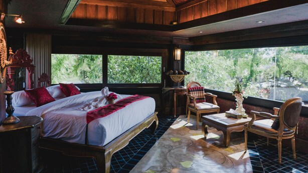 Una habitación con una cama grande, dos sillones, una mesa y vistas al verde exterior.