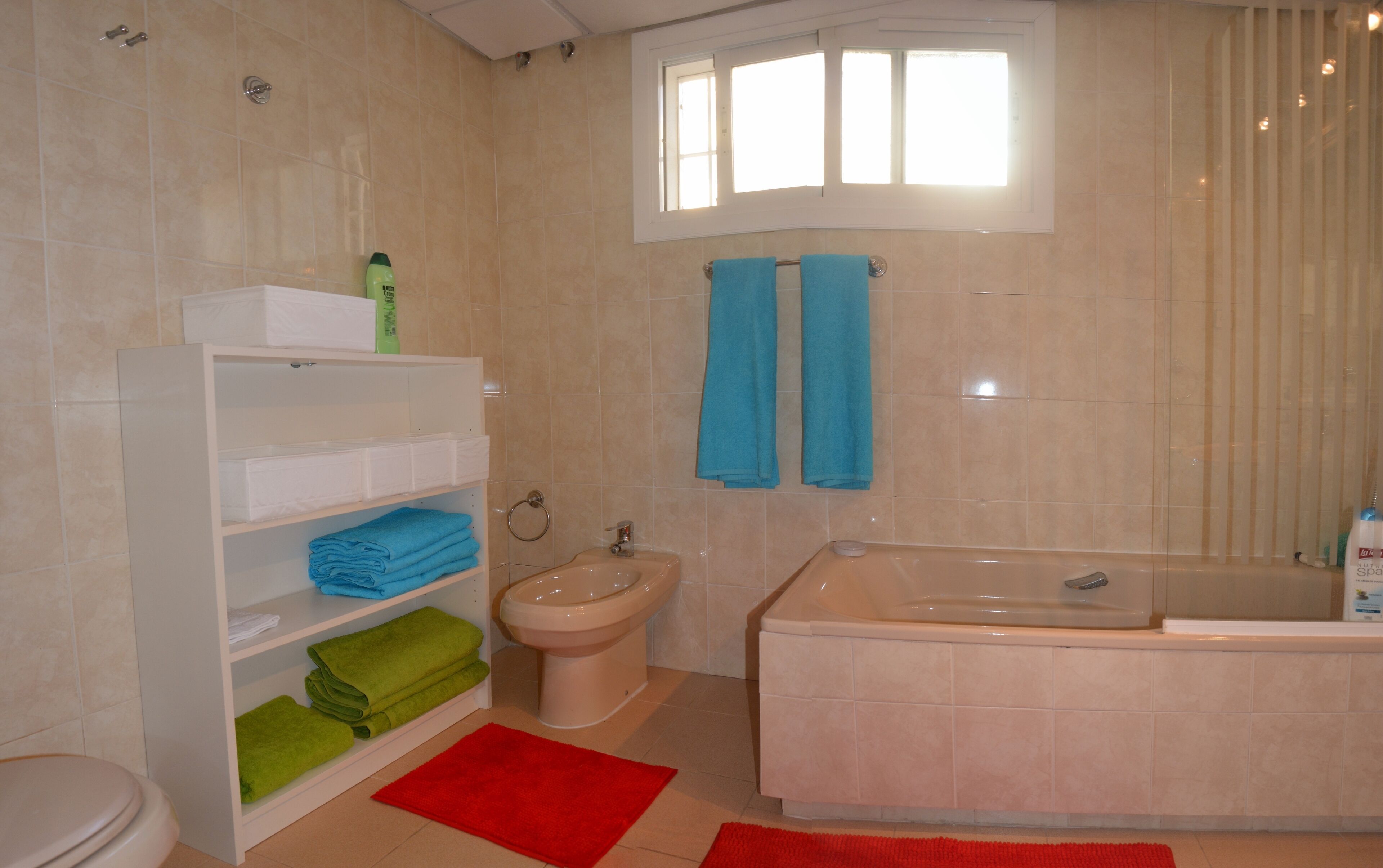 A bathroom with a toilet, sink, bidet, and bathtub. There are shelves with folded towels and a window above the bathtub.