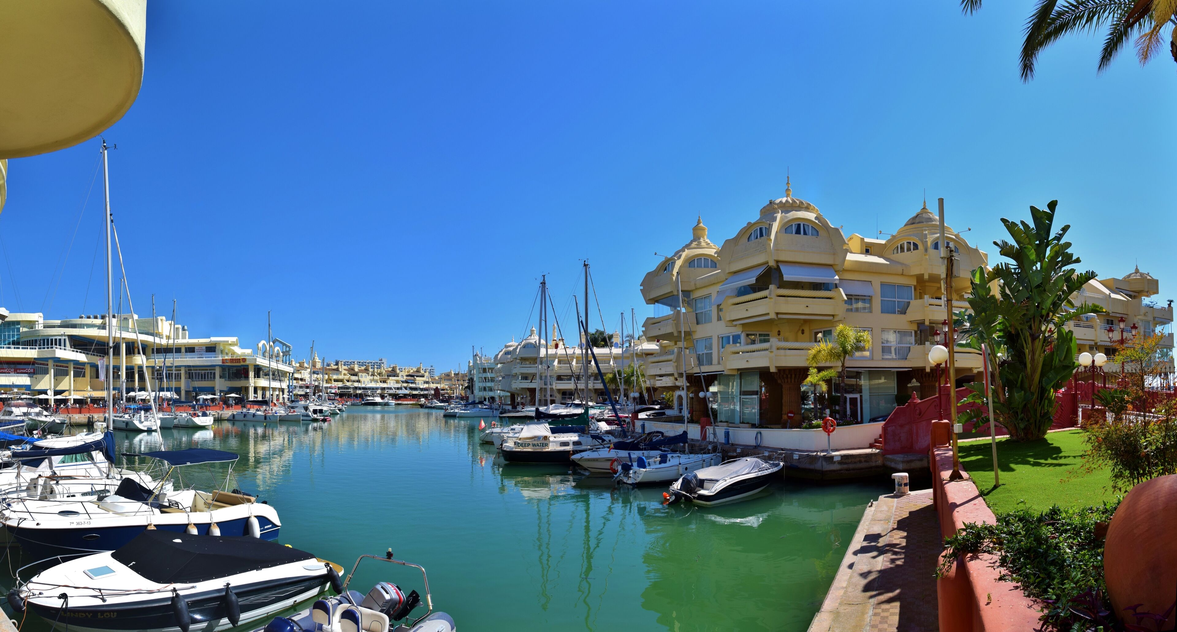A marina with multiple boats docked, a clear blue sky, and a building with a distinctive architectural style.