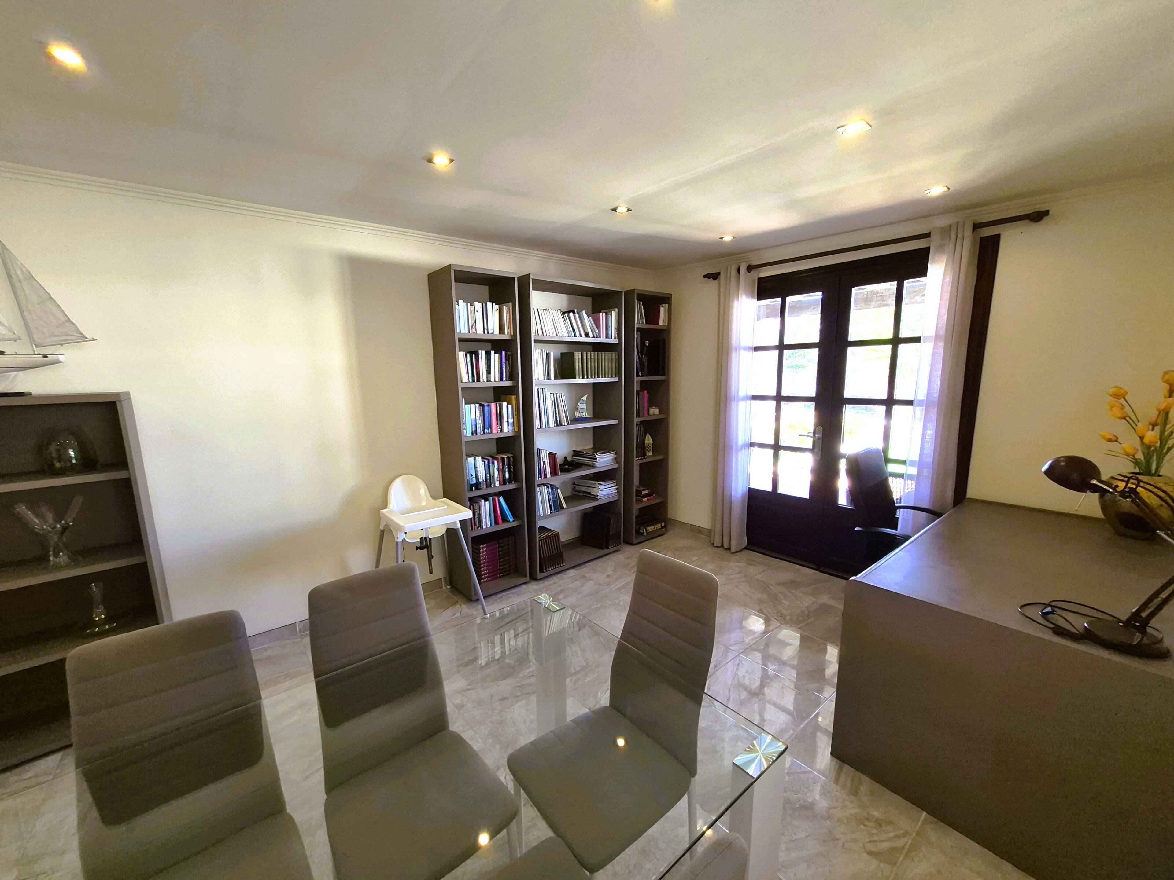 A modern room with a glass table, two chairs, a bookshelf, and a door leading outside.