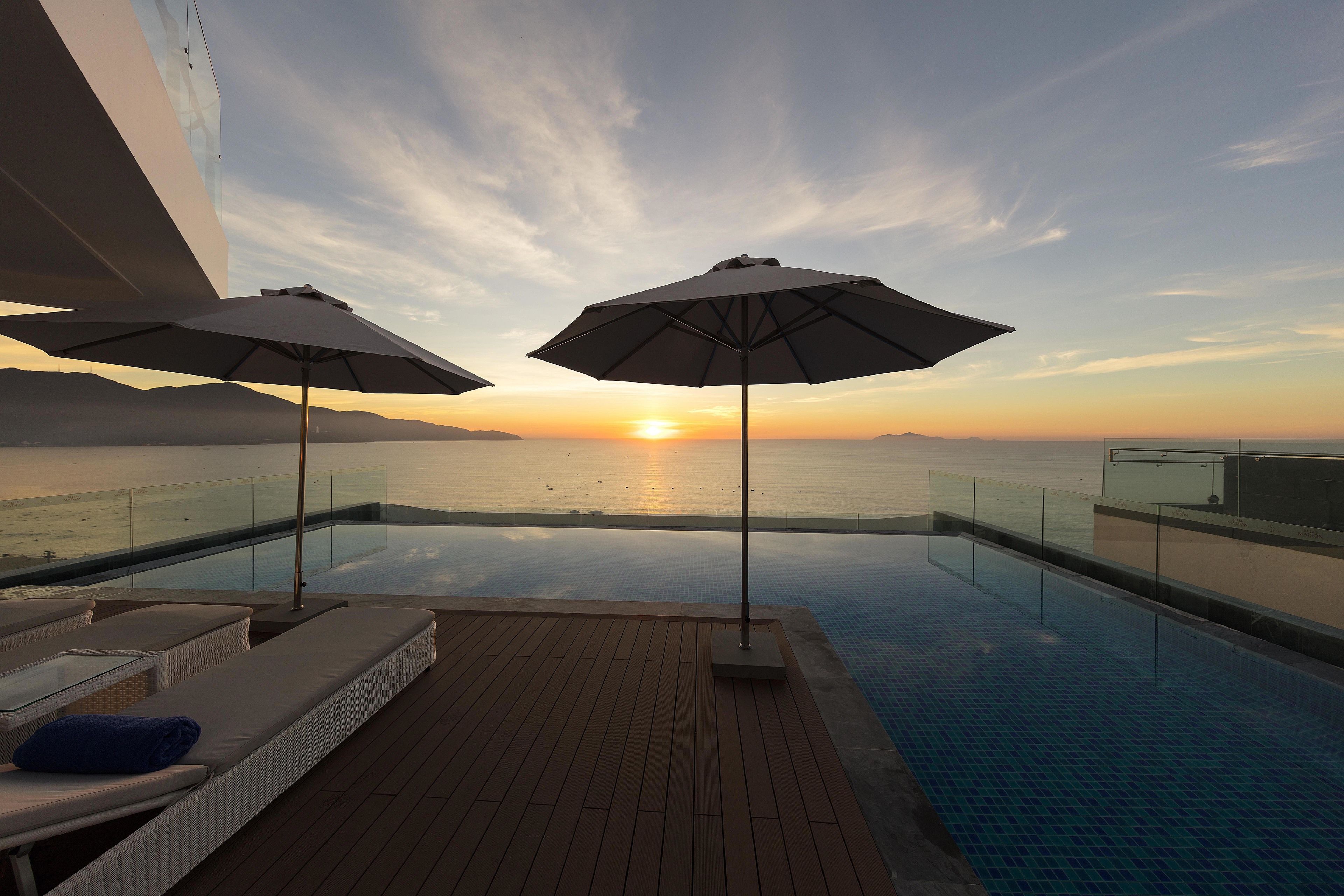 An infinity pool with a clear view of the sunset over the water, two lounge chairs, and umbrellas.