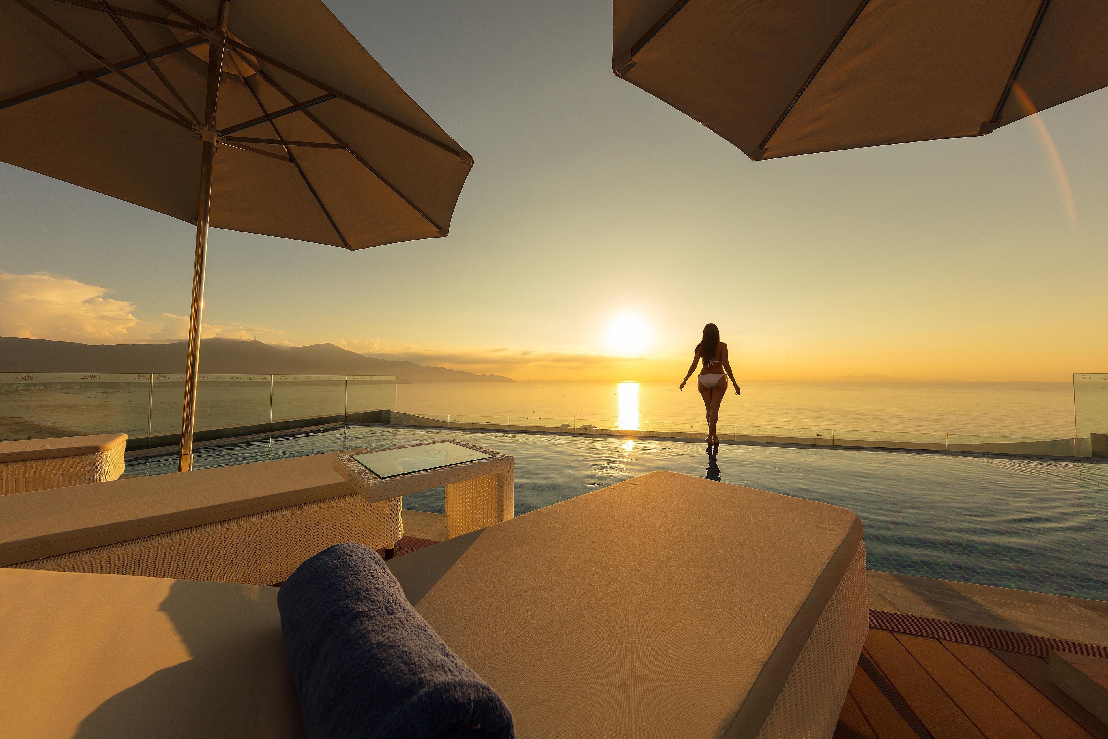 A sunlit infinity pool with lounge chairs and umbrellas, overlooking a sunset over the ocean.