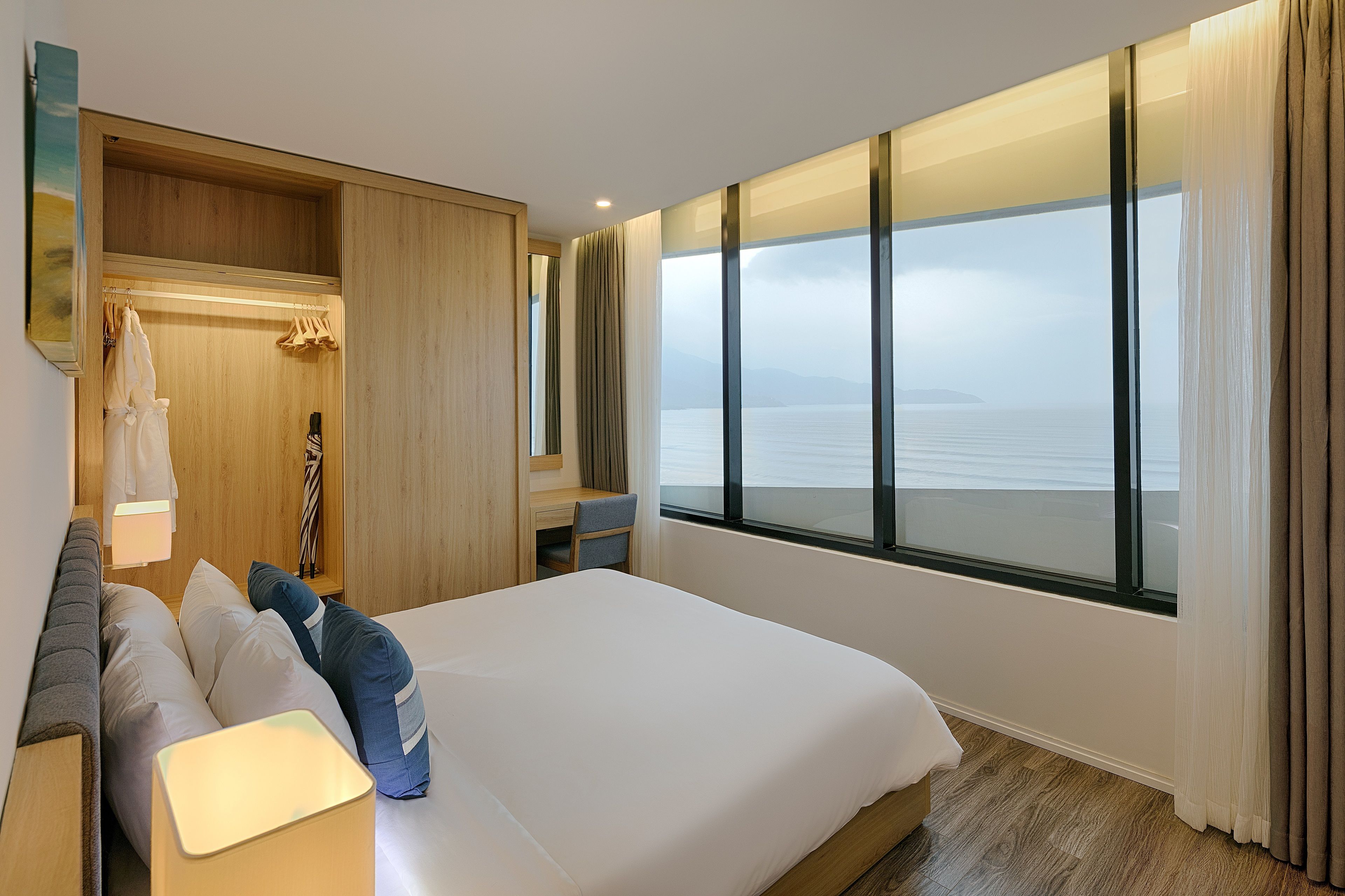 A hotel room with a bed, bedside table, a desk, and a view of the sea.