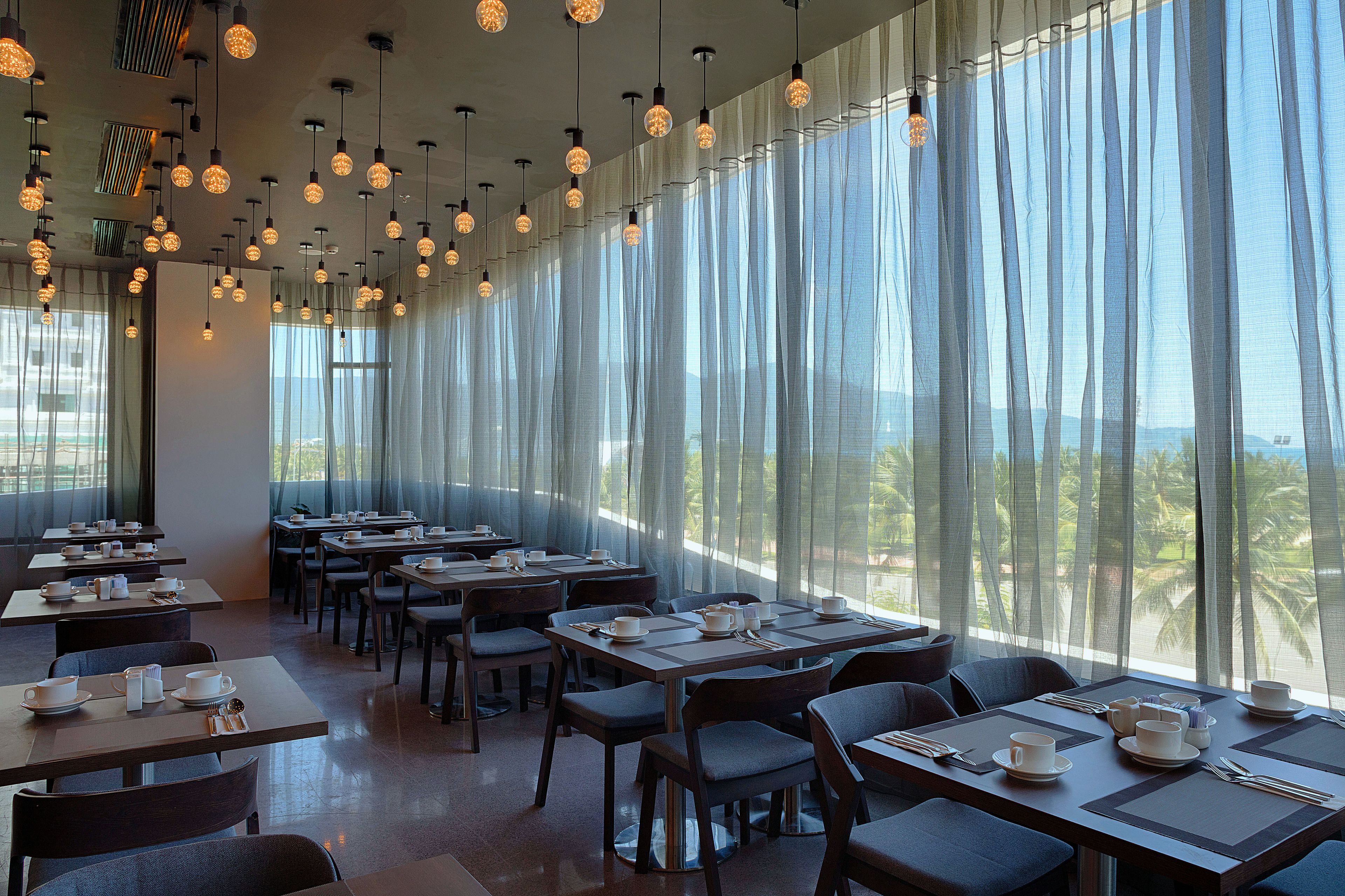 A restaurant with tables set for dining, large windows with sheer curtains, and hanging spherical light fixtures.