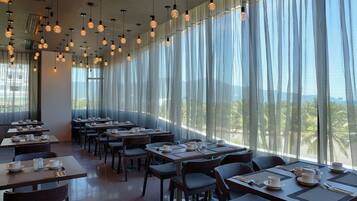 A restaurant with tables set for dining, large windows with sheer curtains, and hanging spherical light fixtures.