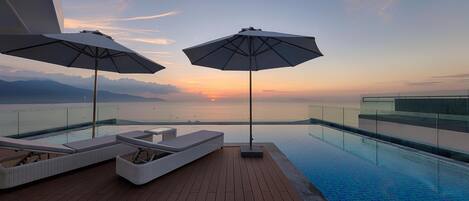 Piscina infinita no terraço com duas cadeiras de lounge brancas e guarda-sóis, com vista para o pôr do sol sobre o oceano.