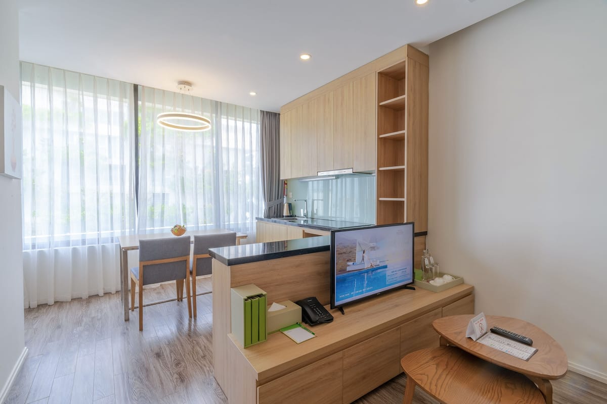 A modern hotel room with a wooden desk, a television, a small table, and a kitchen area.