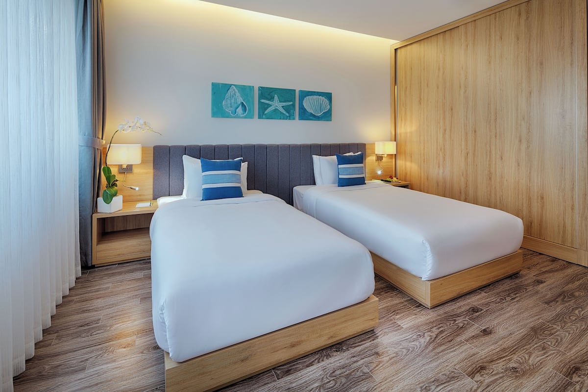 A hotel room with two beds, a wooden wardrobe, a nightstand with a lamp, and wall art.