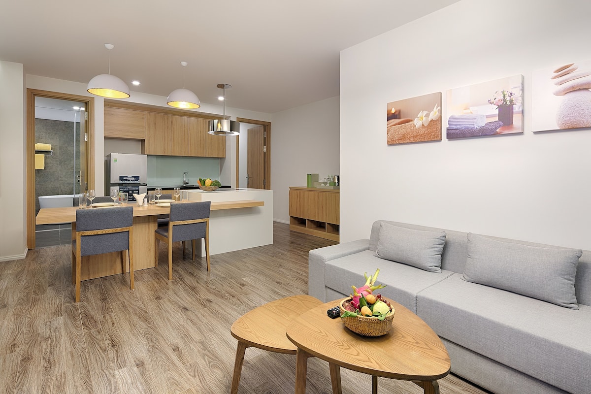 A modern living room with a grey sofa, wooden dining table, and a kitchen area with wooden cabinets and pendant lighting.