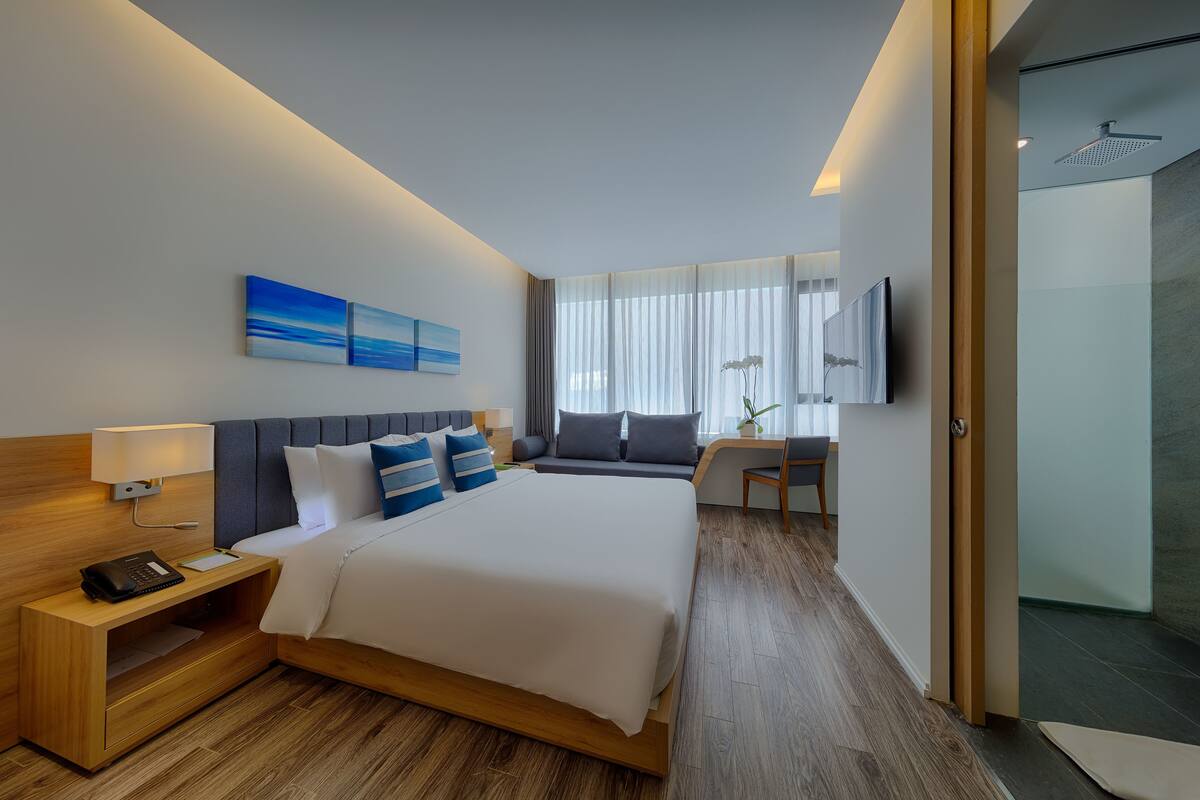 A modern hotel room with a large bed, bedside table, lamp, and a desk with a chair.