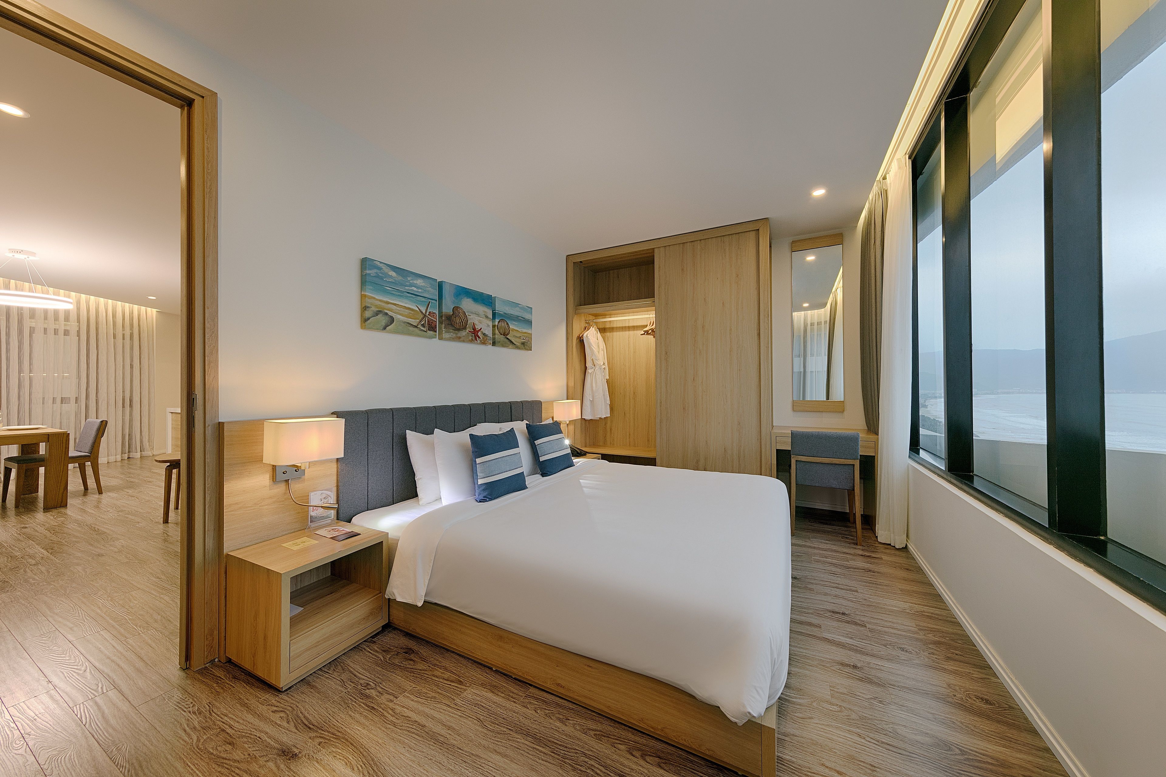 A modern hotel room with a large bed, wooden headboard, bedside tables, a desk, and a view of the sea.