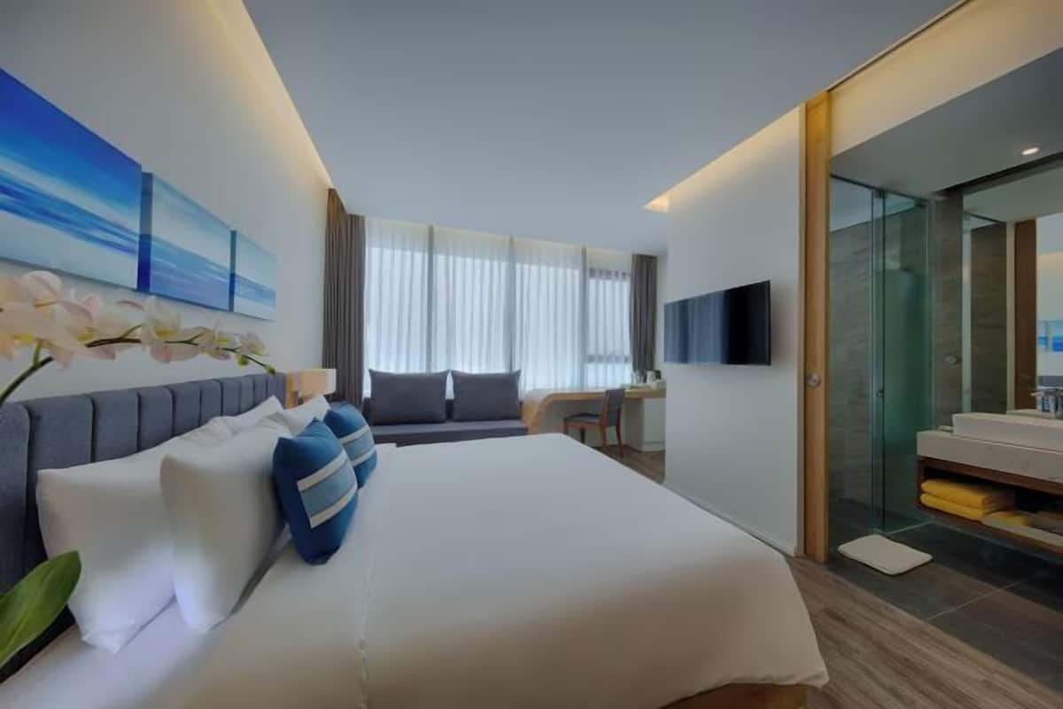 A modern hotel room with a large bed, a desk, a TV, and a glass shower area.
