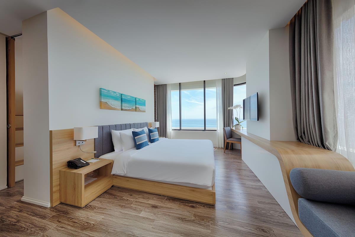 A modern hotel room with a large bed, bedside table, lamp, and a view of the ocean.