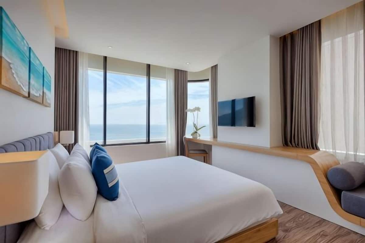 A hotel room with a bed, a desk, a TV, and a view of the sea.