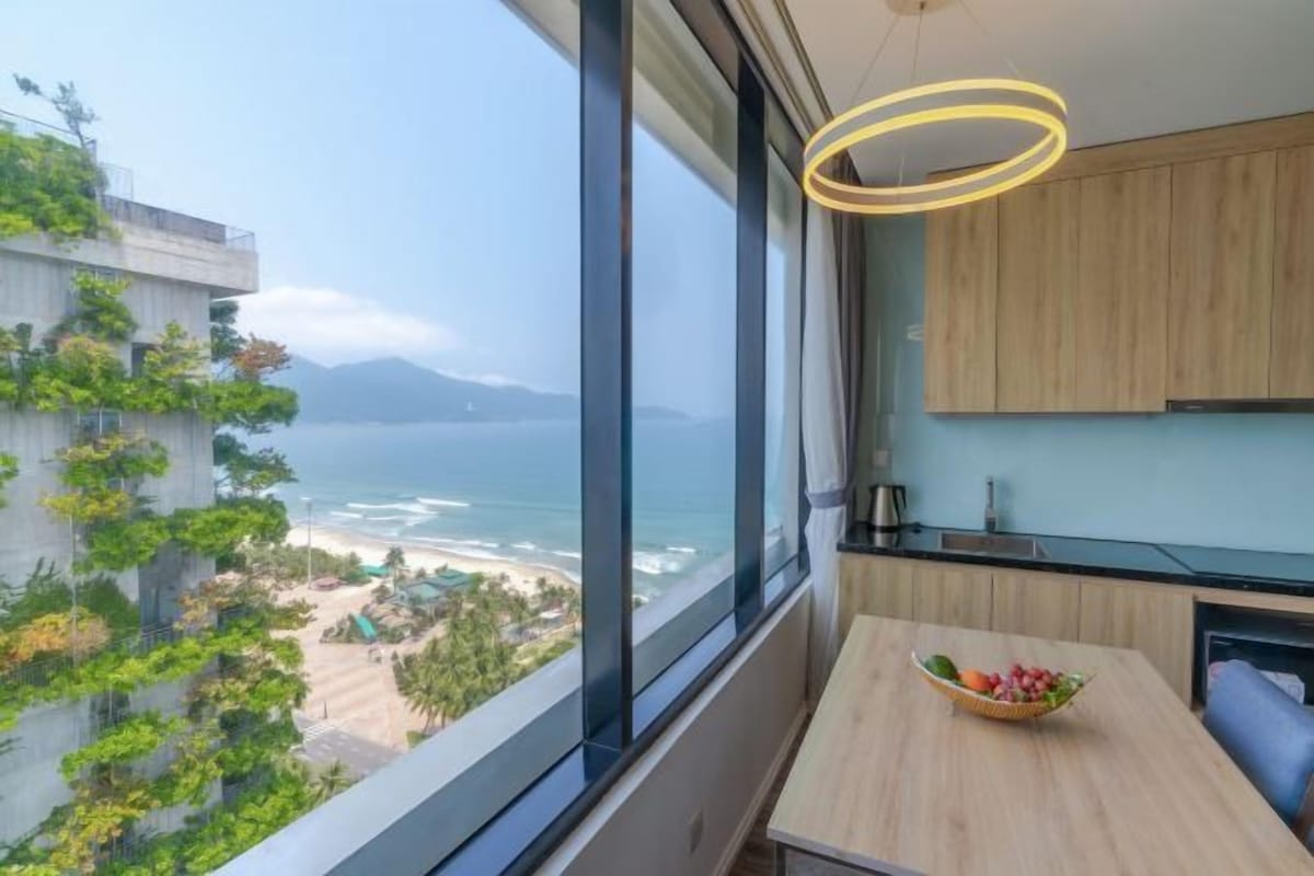 Une cuisine moderne avec vue sur l’océan, une table à manger agrémentée d’un panier de fruits et une grande fenêtre.