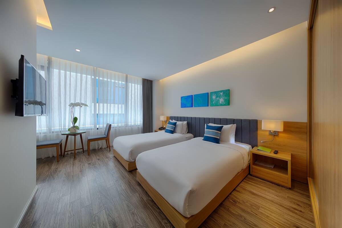 A hotel room with two beds, a desk with a chair, a TV, and a nightstand with a lamp.