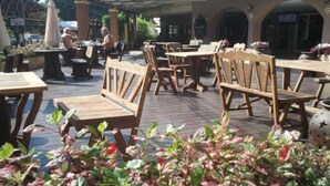 An outdoor cafe area with wooden tables and chairs, surrounded by potted plants and a brick wall.