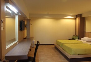 Habitación de hotel con un escritorio de madera alargado, dos camas individuales con ropa de cama verde, un televisor de pantalla plana montado en la pared y una pequeña zona de estar con una silla.