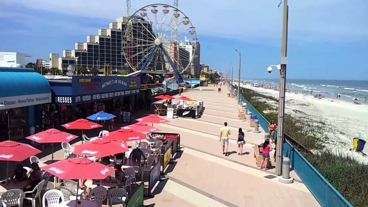A boardwalk with outdoor seating, a Ferris wheel, and a beach in the background.