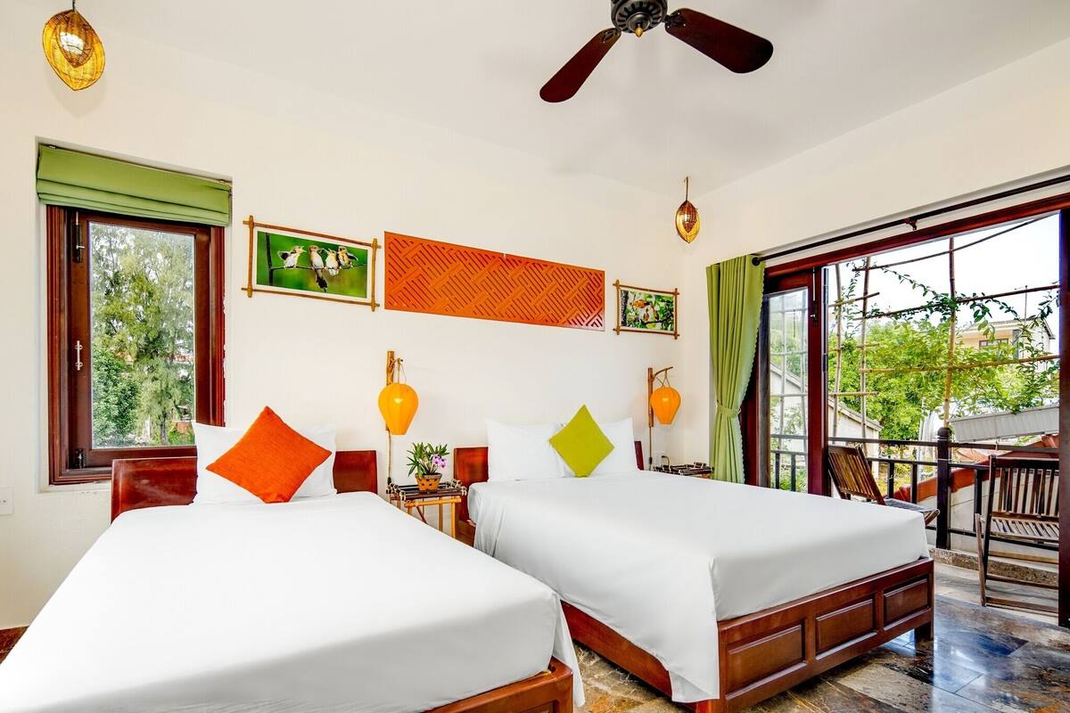 Une chambre d’hôtel avec deux lits, un ventilateur de plafond, de grandes fenêtres et un balcon aménagé avec du mobilier d’extérieur.