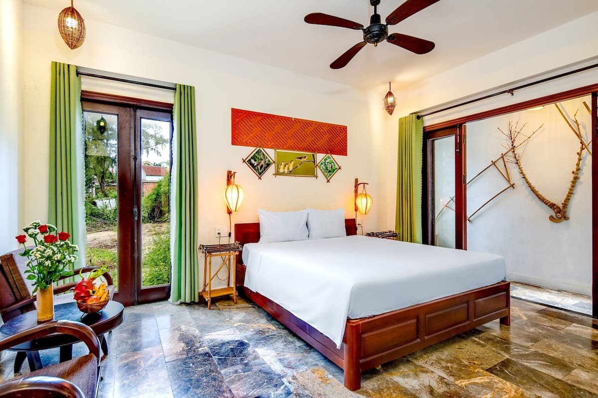 Une chambre d’hôtel avec un lit, une table à manger avec des chaises, un vase de fleurs et une vue sur l’extérieur par la porte.