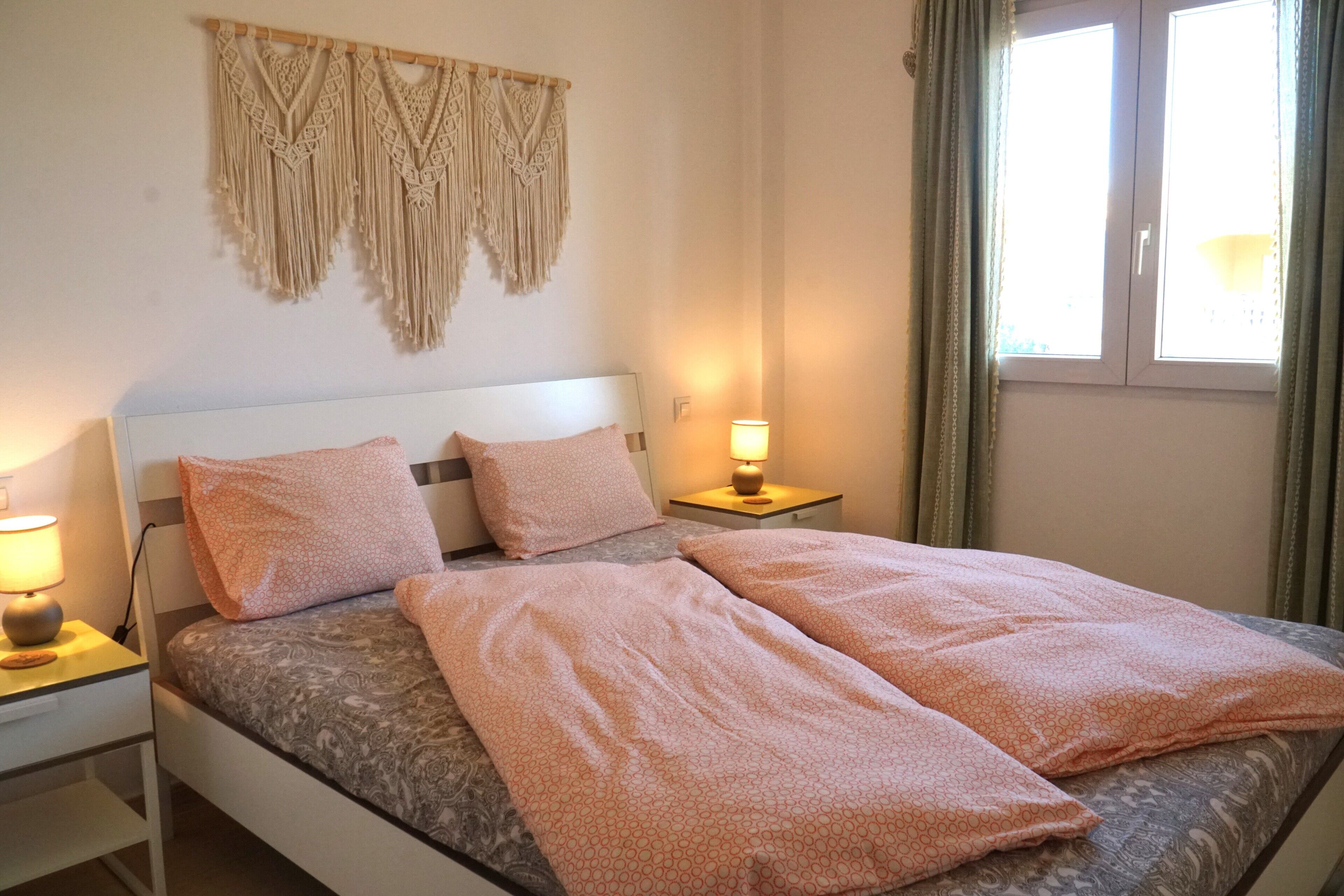 Un lit bien fait, avec deux oreillers et une couverture, une lampe de chevet, une décoration murale en macramé et une fenêtre avec des rideaux.