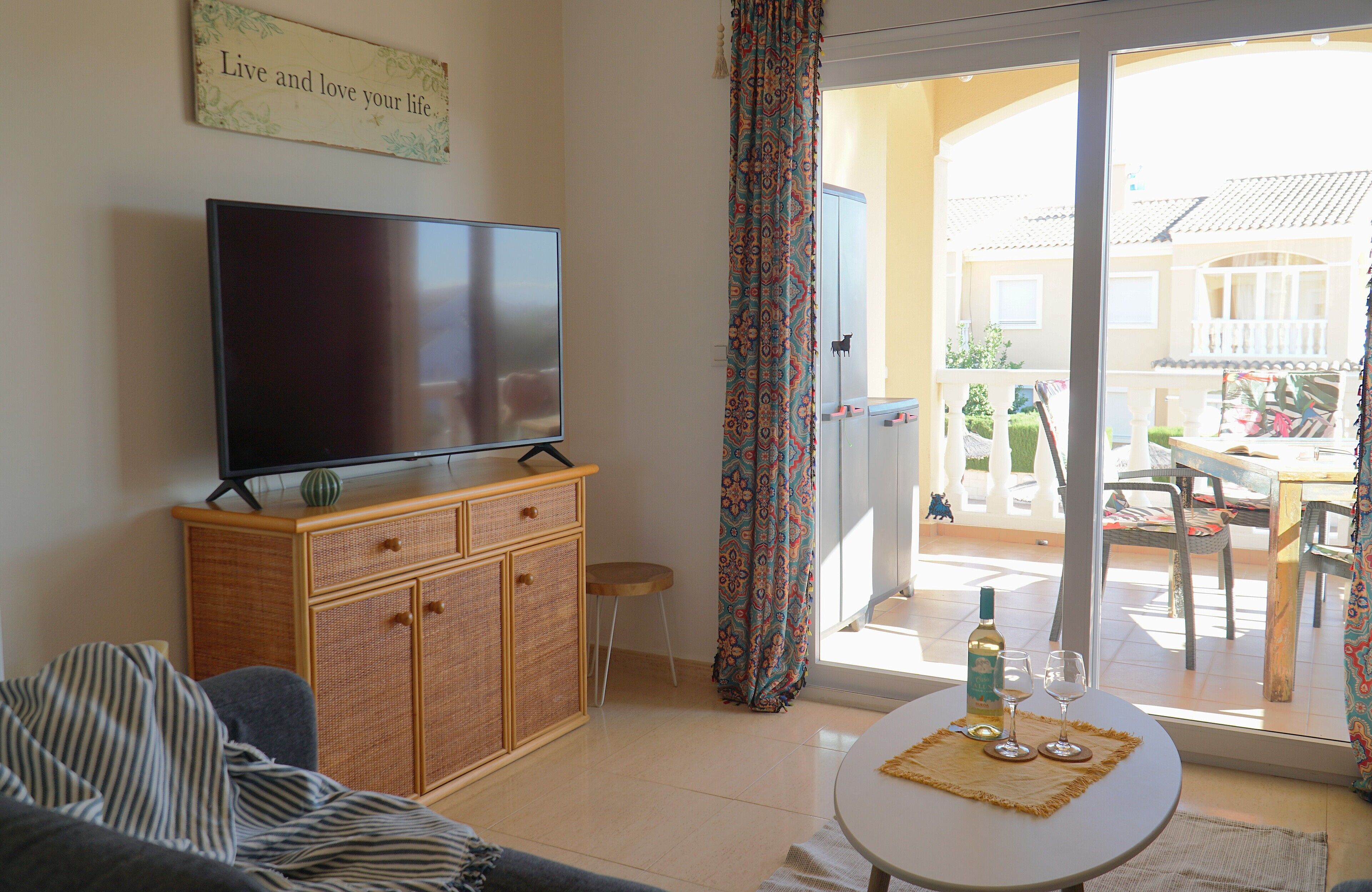 A living room with a television, a sofa, a small table with a bottle and glasses, and a sliding glass door leading to an outdoor patio.