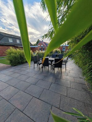 A patio with a table and chairs, surrounded by greenery and a brick house in the background.