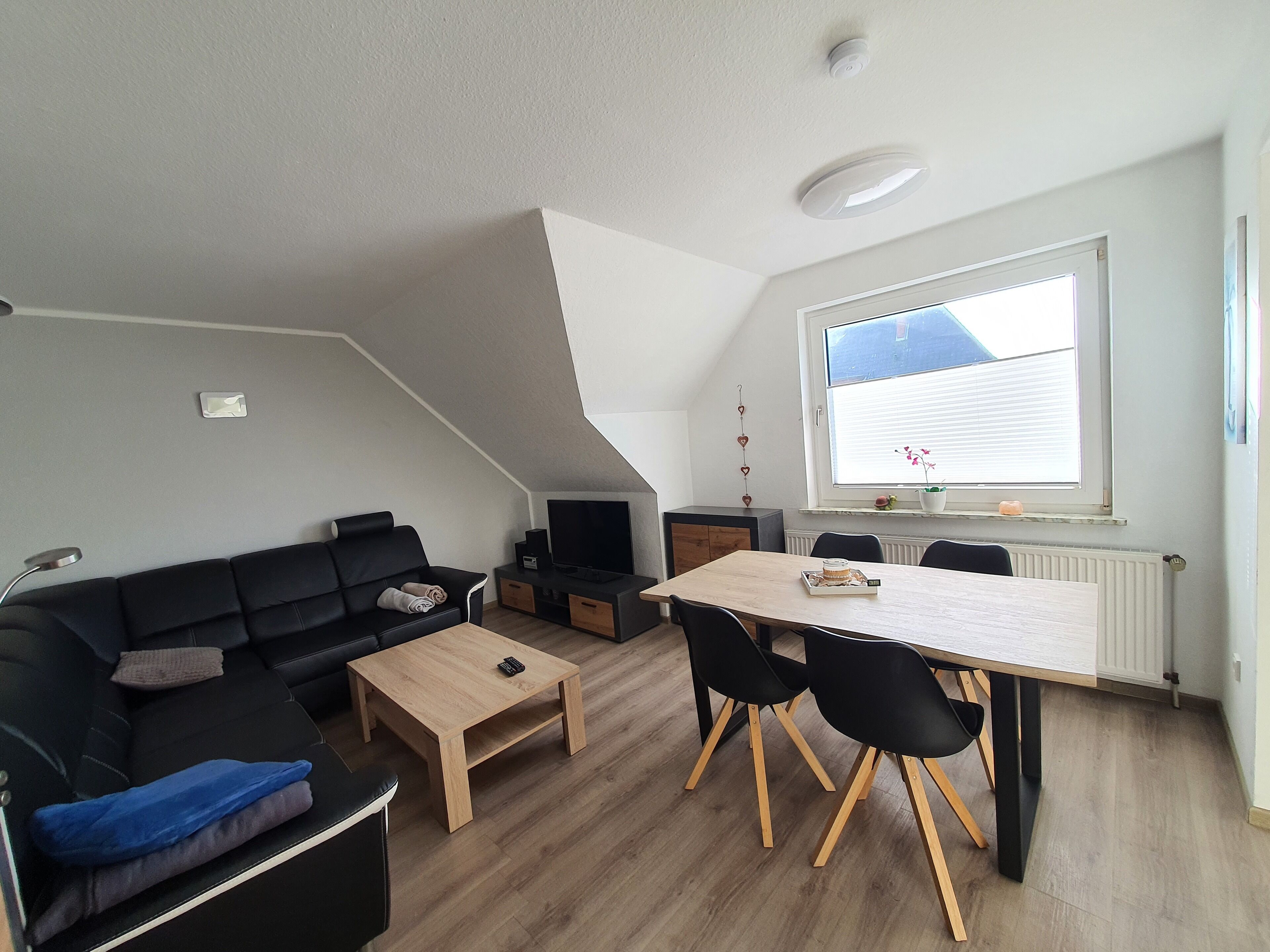 Un salon moderne avec un canapé noir, une table basse en bois, un téléviseur et un coin repas comprenant une table et des chaises.